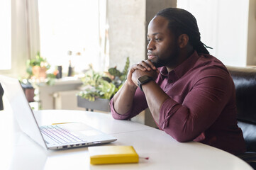 Focused and concerned black man in smart casual shirt looks at laptop screen, serious...