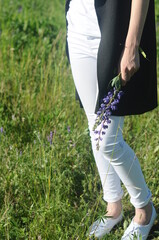 young caucasian woman in white trousers and a black jacket side view holds a bouquet of lupines in her hand on the summer green field background closeup. Selective focus