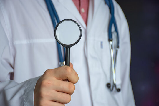 Doctor with a magnifying glass in his hands against the background of a white coat. Concept photo for diagnosing and searching for diseases