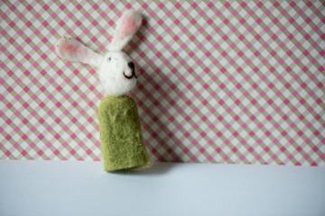 a white rabbit in a green cloak finger puppet posing against a pink and green plaid background