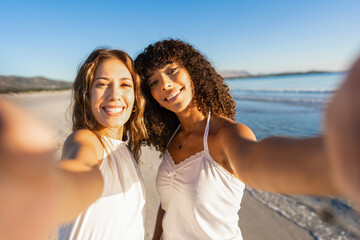 Two beautiful mixed race girl holding smartphone making a self portrait on beach smiling at camera wearing boho white dress at sunset or dawn. Gay couple of young millennial women in tropical vacation