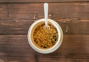 Instant noodles with a sticking out plastic fork on a wooden table. Top view close up