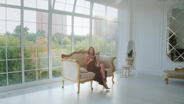 Gorgeous brunette woman wearing black dress sitting on a couch in front of a big panoramic window filled with the sunshine. High quality 4k footage