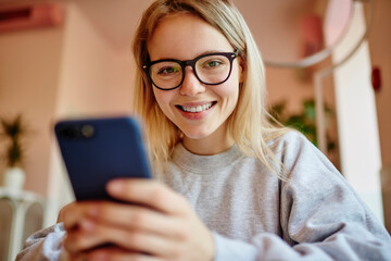Portrait of cheerful millennial blogger holding digital smartphone gadget and smiling at camera,...