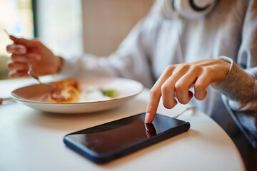 Unrecognizable female tapping on touchscreen of modern cellphone gadget during healthy breakfast in cafe interior,selective focus on digital smartphone technology used for online browsing in cafeteria
