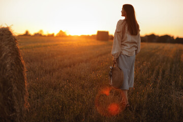 Fototapeta premium young woman in the beautiful light of the summer sunset in a field is walking near the straw bales. beautiful romantic girl with long hair outdoors in field.
