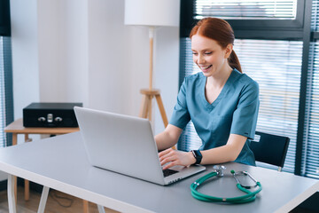 Portrait of cheerful smiling young female doctor in blue green medical uniform typing on laptop computer looking on screen, sitting at desk near window in modern office of medic clinic.