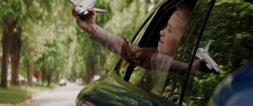 CU Cute little dreamer child girl sticking head out of car window and flying her toy spaceship while riding through neighborhood. Shot with 2x anamorphic lens
