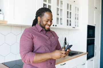 Hilarious guy with dreadlocks using smartphone for communication and messaging online, African-American man enjoys chatting in social networks, looks at the screen and smiles standing in the kitchen