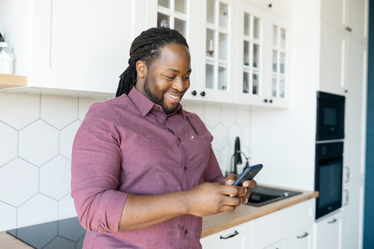 Hilarious guy with dreadlocks using smartphone for communication and messaging online, African-American man enjoys chatting in social networks, looks at the screen and smiles standing in the kitchen