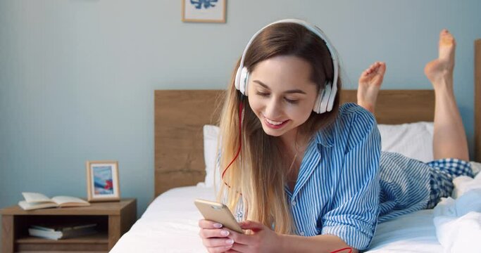 Smiling woman wearing headphones looking at the smartphone while laying on white bed. Happy young woman using gadgets at home. Beautiful girl listening music and surfing the net with pleasure smile