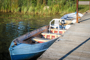 Metal row boats - one with plastic chair in it- pulled up to pier with mossy weedy pond bank behind.