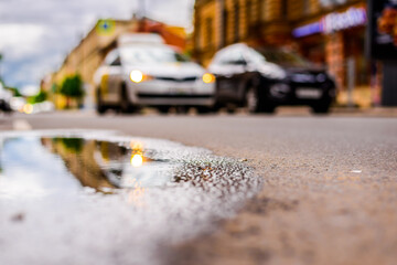 Sunny day after rain in the city, parked cars near the store and a passing car. Close up view from the level of the puddle on the pavement
