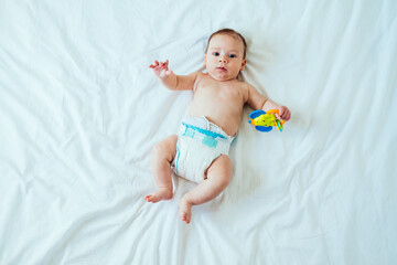 Portrait of Cute Baby Looking at Camera and Holding Rattle Toy in Hand on White Sheets with Copy Space .
Top view of adorable newborn baby in diaper lying on bed and holding rattle toy. - obrazy, fototapety, plakaty