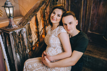 Stylish man hugs his beloved pregnant woman in a chic lace dress, sitting on the stairs in a vintage interior.