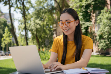 Beautiful asian woman wearing stylish eyeglasses using laptop computer, typing, planning freelance project, working outdoors. Young styling student studying, exam preparation, online education