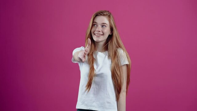 Welcome. The kind, welcoming young woman extends her hand, delighted with knowledge. Freckled teenage girl in a white T-shirt, isolated on a pink background. The concept of people's lifestyle