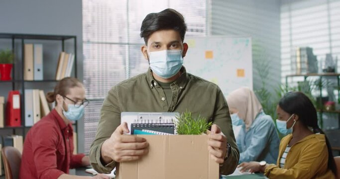 Portrait of young handsome Hindu man employee in medical mask standing in cabinet holding carton box with his goods, fired from work. Mixed-race workers working behind at office, occupation concept