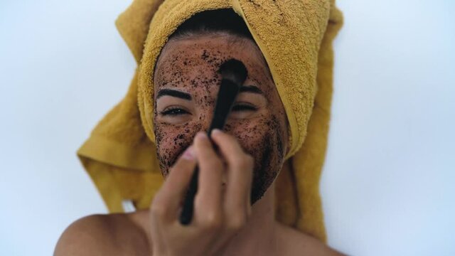 Young smiling woman applying coffee scrub mask with brush on face - Happy girl having skin care spa day at home - Healthy alternative natural exfoliation treatment and youth people lifestyle concept
