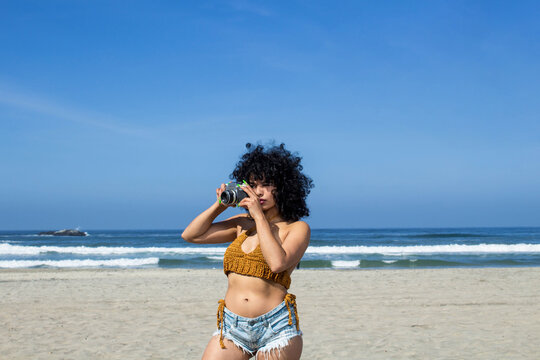 beautiful afro woman taking pictures on the beach with old camera