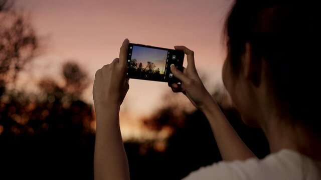 Young happy asian girl hold mobile phone relax cheerful take photo peaceful picture of sunset at forest tree in park. Beautiful nature sunset view, Travel holiday vacation with smartphone concept.