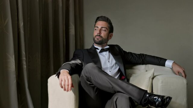 Image Of An Elegant Young Man In Suit. He Is Sitting On The Couch. His Hand Touches His Chin. He Thought.