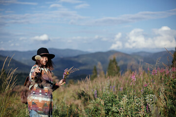 Happy woman traveler with backpack and hat gathering wildflowers on mountain hills and relaxing