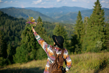 Woman traveler with backpack and hat holding bouquet of wildflowers on background of mountain hills