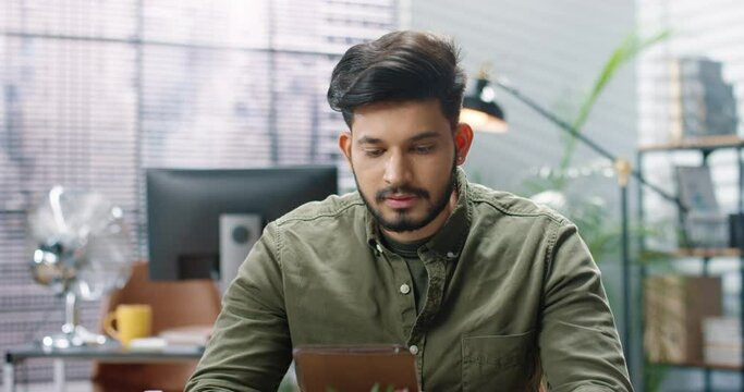 Close up portrait of Hindu joyful young handsome male worker texting on tablet browsing online while sitting at workplace in cabinet looking at camera and smiling Office concept successful businessman