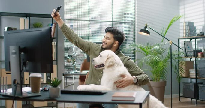 Happy joyful Hindu male sitting in cabinet at work making selfie photo with his cute dog pet in good mood. Handsome cheerful bearded guy at office taking pictures with animal on smartphone camera