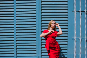 Fototapeta premium A young woman stands near a wall with blue shutters. Bright sunny day, hard light.