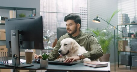 Portrait of joyful Hindu handsome man sitting in cabinet at work with his cute dog pet in good mood typing on computer browsing online. Positive emotions, pet concept. Work in office