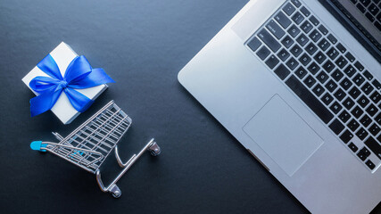 Web shopping. Laptop computer, shopping trolley and white gift with blue ribbon on dark background. Ecommerce and customer experience concept. Internet purchase, online shop.