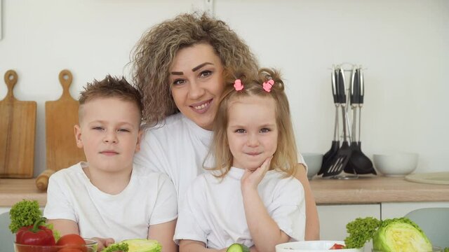 Children with mom sitting at a table in a bright kitchen. Fresh vegetables are on the table. Healthy Eating. Children's menu. The family is dressed in the same basic clothes of white and blue colors
