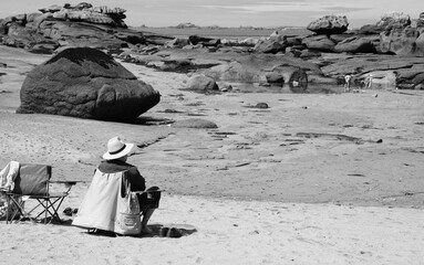 Senior man (unrecognizable; back view) relaxing at Pink Granite Coast beach and regard his grandchildren looking for clams. Brittany, France. Family summer vacation background. Black white photo