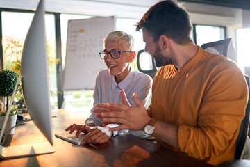 A young businessman and elderly female colleague have a conversation about a computer screen content watching together at the desk at workplace. Business, office, job - Powered by Adobe