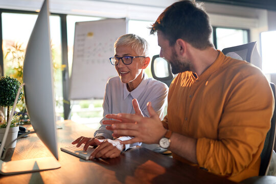 A Young Businessman And Elderly Female Colleague Have A Conversation About A Computer Screen Content Watching Together At The Desk At Workplace. Business, Office, Job