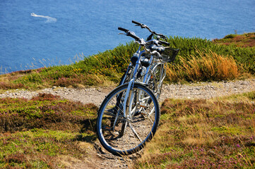 Fototapeta na wymiar Two bicycles on the hill over the sea in sunny day and a boat sailing at background. Breton coast near Cap de la Chevre. Brittany, France. Healthy lifestyle. Active summer vacation background.