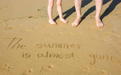 A sentence written on the sand of a beach: THE SUMMER IS ALMOST GONE, footprints and male and...