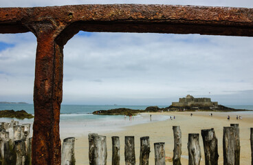 A view on Fort National (on a tidal island) and the Saint-Malo beach (with resting and walking people) during the low tide. Brittany, France. Selective focus on the rusty fence.