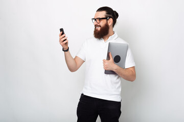Photo of young man with beard in white t-shirt using smartphone and holding laptop