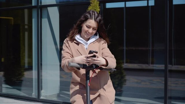 A young student girl in a sweater, jeans and a coat stands with an electric scooter near a stylish modern building and types in a smartphone. Front view
