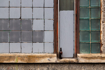 Old wall with a window made of square gray and glass tiles. Empty bottle on the windowsill. Rough...