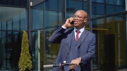 Young male office worker talking on the phone.African american man in a classic blue business suit stands with an electric scooter on the street near a modern mirror building and talks on the phone