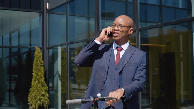 Young male office worker talking on the phone.African american man in a classic blue business suit stands with an electric scooter on the street near a modern mirror building and talks on the phone - Powered by Adobe