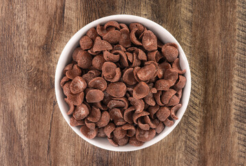 Top view of bowl with sweet chocolate cereal flakes for breakfast on white wooden background