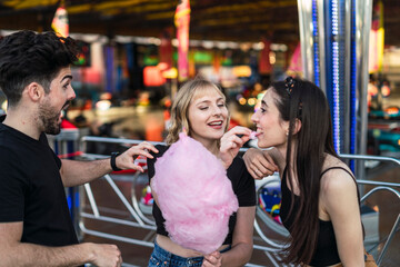Dos chicas y un chico amigos sonriendo y compartiendo un algodón de azúcar rosa en zona de atracciones de una feria