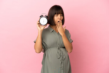 Fototapeta premium Woman over isolated background pregnant and holding clock while yawning