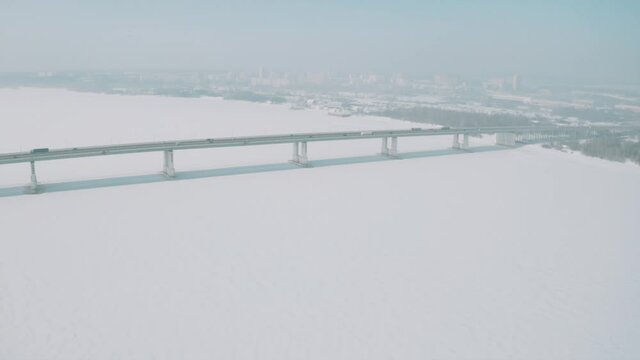 Aerial view of a long bridge above snow and ice covered river between two parts of a city. Clip. Winter landscape with a wide white river and a bridge with driving cars.