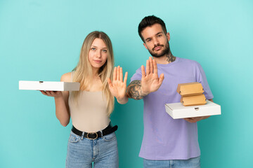 Couple holding pizzas and burgers over isolated blue background making stop gesture denying a situation that thinks wrong